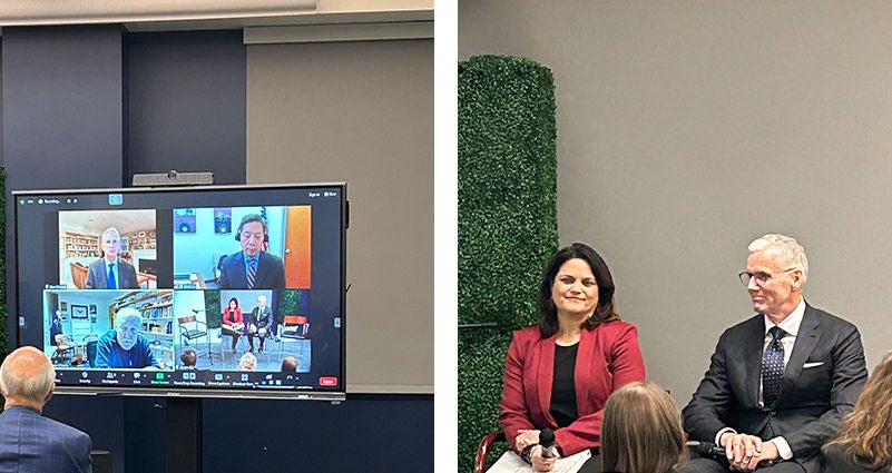 A hybrid business event shows a large screen displaying a multi-person video conference on the left side. Several professionals appear in separate video windows, participating remotely from office and home environments. On the right side, two speakers sit on a stage in front of a gray wall and green decorative panel, addressing an in-person audience. The scene highlights modern corporate communication that blends virtual and in-person participation.