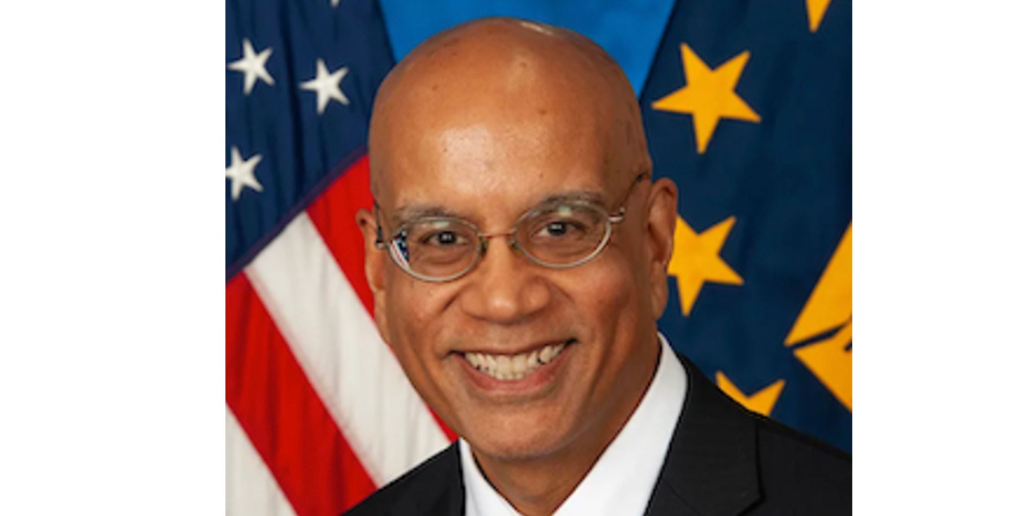 A formal portrait featuring a person in a suit against a backdrop of the American flag and a blue banner with yellow stars. The setting suggests an official or governmental context. The image conveys a professional and patriotic tone.
