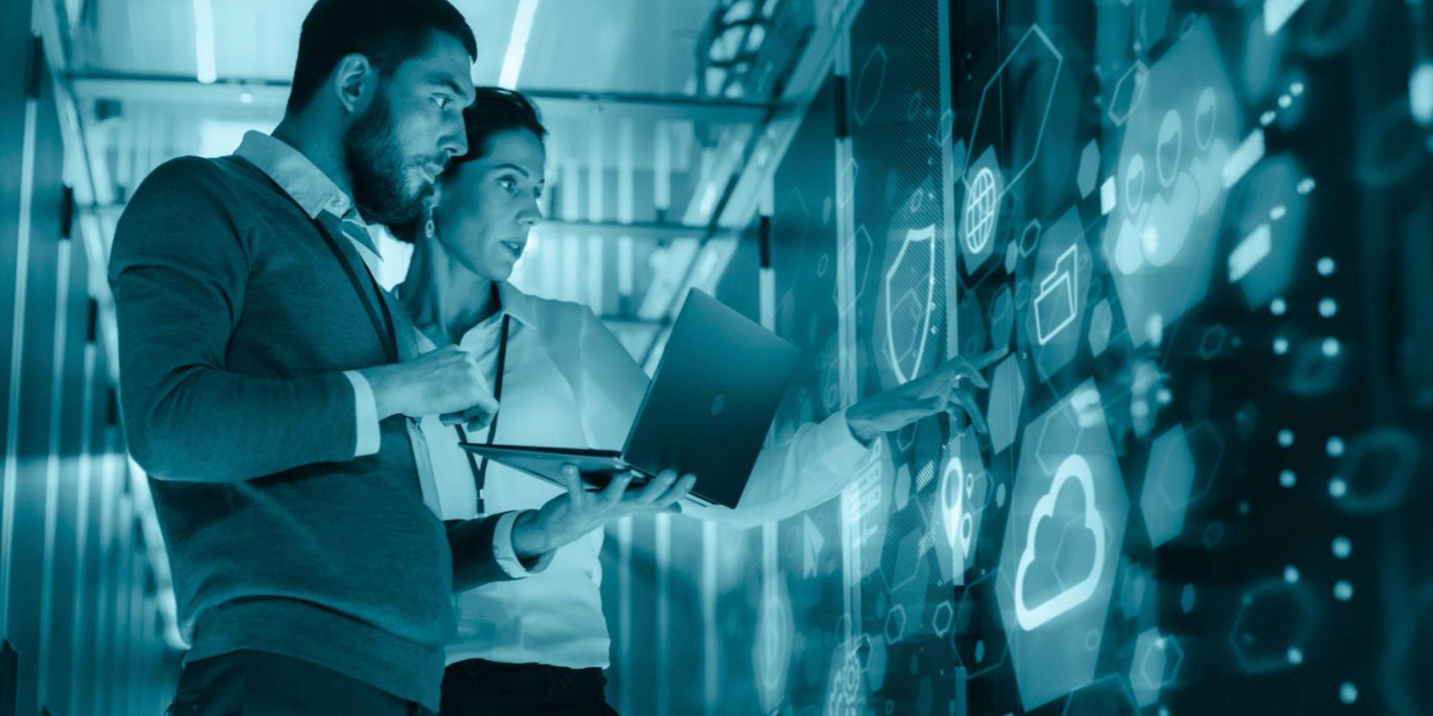 Two professionals collaborate in a high-tech data center, interacting with a large digital touchscreen displaying cloud and network icons. The setting features a modern, blue-toned palette with illuminated technology visuals. One person holds a laptop while the other points at the screen, emphasizing teamwork and digital innovation. The environment suggests advanced IT infrastructure and cloud computing concepts.