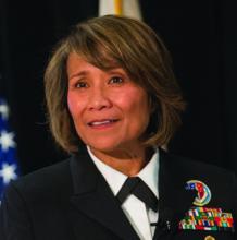 A female naval officer is shown in a formal dress uniform, speaking or posing in front of a dark backdrop. Colorful service ribbons and an insignia are visible on her jacket, indicating military honors. The background includes a blurred flag, suggesting an official or ceremonial setting. Her expression appears calm and composed, contributing to a respectful and professional atmosphere.