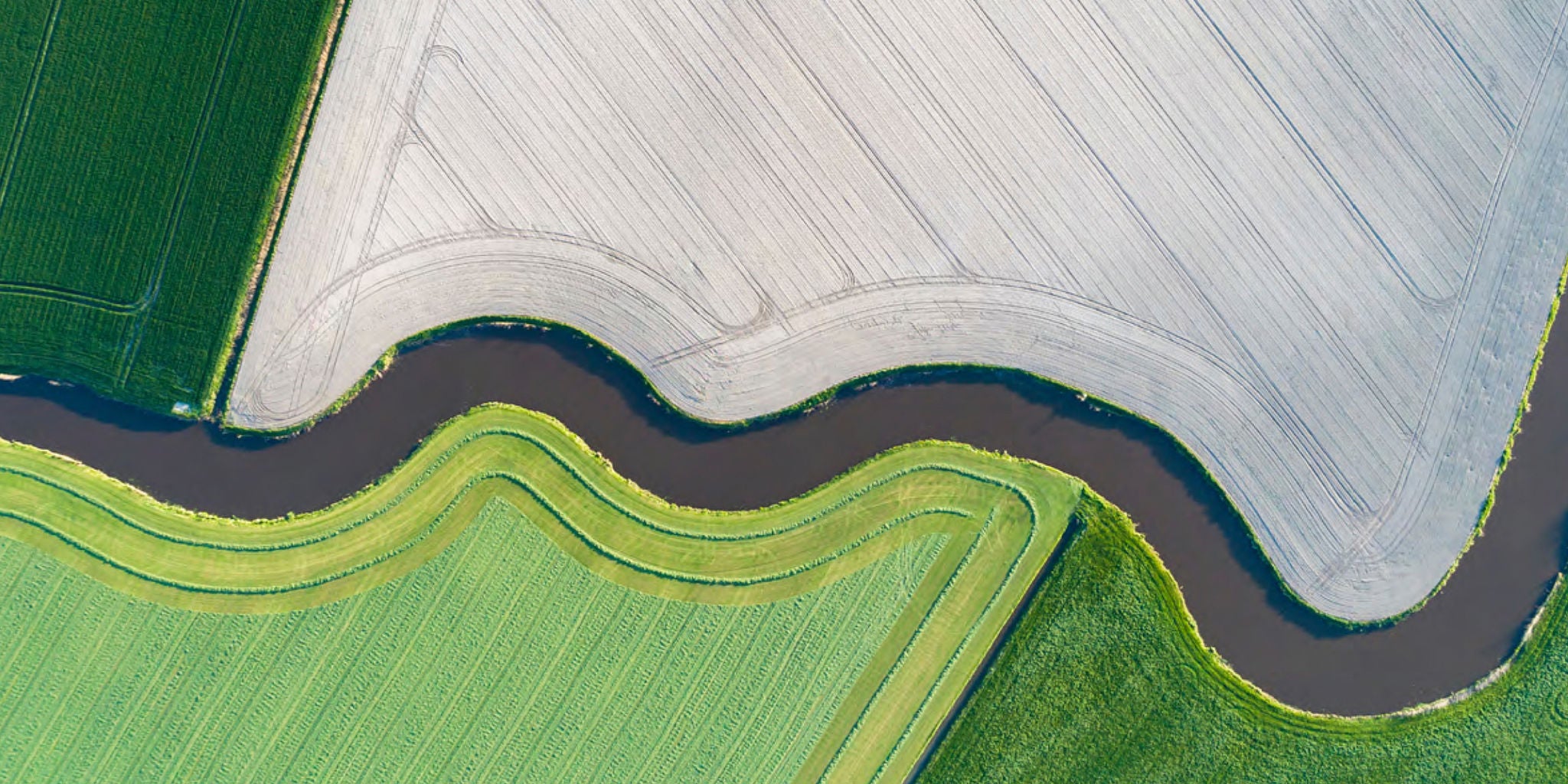 This aerial photograph captures a striking landscape of cultivated farmland divided by a winding waterway. The fields display contrasting colors and textures, with one side appearing freshly plowed and the other lush and green. The curving lines and geometric patterns create a visually dynamic composition. No visible text, numbers, or identifiable brands are present in the image.