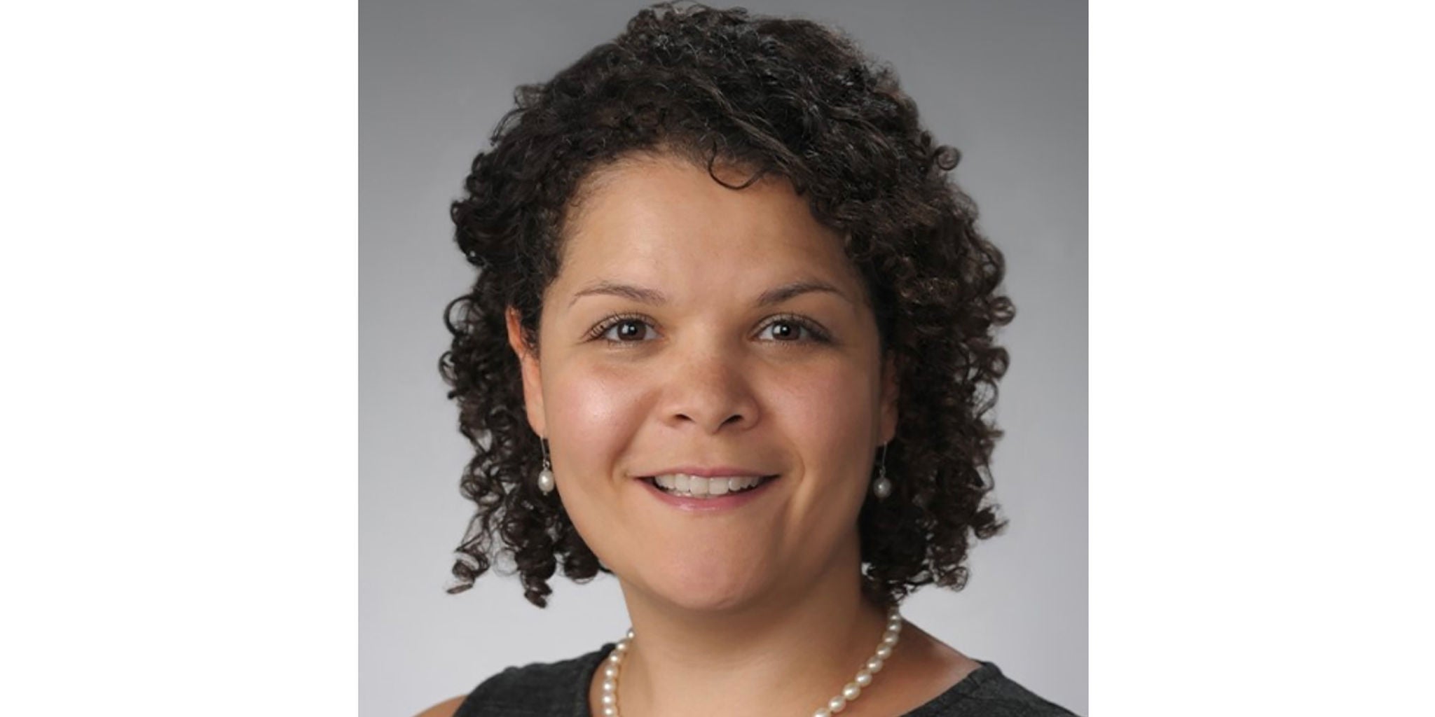 A professional portrait featuring a person wearing a black top and a pearl necklace. The background is a neutral gray, creating a formal and polished look. The focus is on the attire and accessories, emphasizing elegance and professionalism.