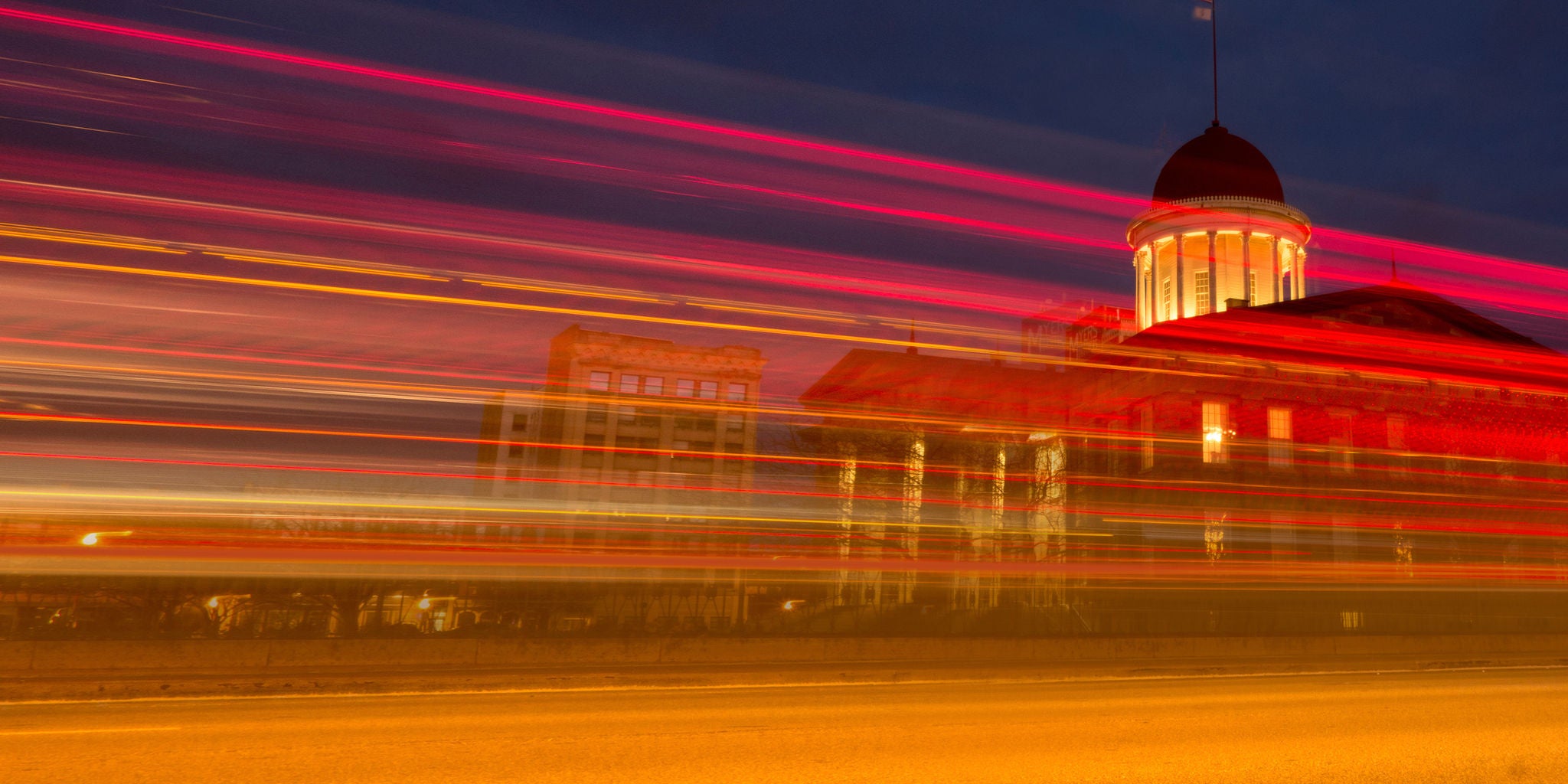 A historic domed building is illuminated against a dark evening sky. Bright red and yellow light trails streak horizontally across the scene, suggesting moving vehicles. The long exposure effect creates a dynamic, energetic visual contrast with the still architecture. The overall palette features warm tones and deep blues. No visible text or numbers are present in the image.