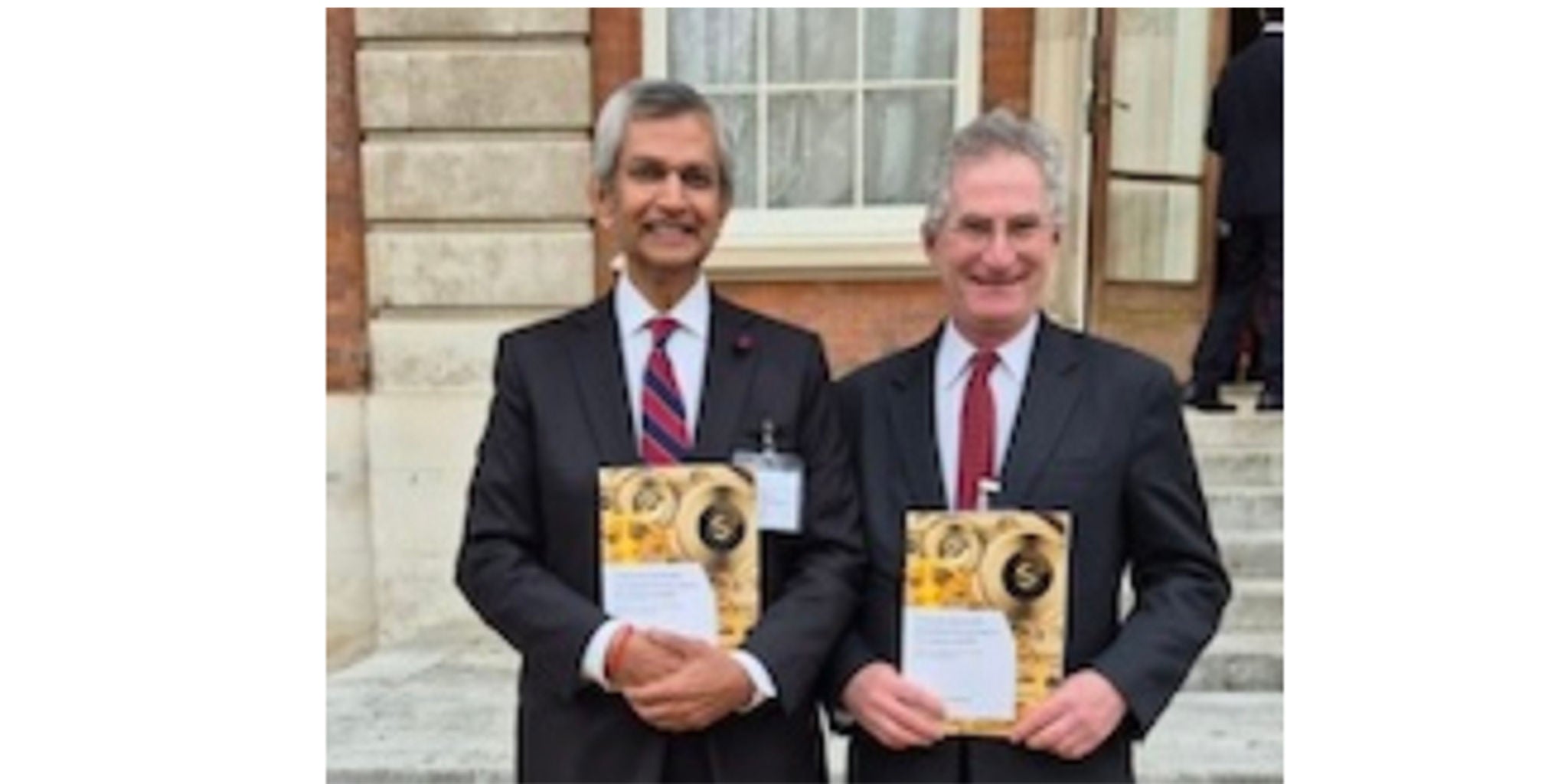 Two individuals dressed in formal suits are standing outdoors, holding certificates with visible gold and white designs. The setting appears to be in front of a historic building with stone steps and large windows. The certificates feature a circular gold emblem and text, suggesting an award or recognition event.