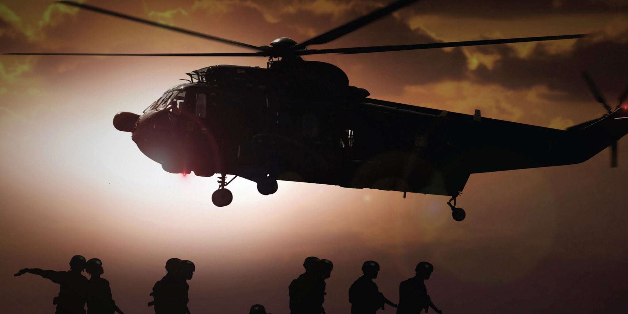 A large military helicopter is silhouetted against a dramatic sunset sky, preparing to land or take off. Several soldiers are visible in the foreground, walking or preparing for action, their outlines sharply defined by the backlighting. The scene conveys a sense of urgency and teamwork, with warm, muted tones dominating the palette. No visible text or numbers are present in the image.
