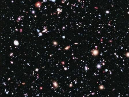 A vast field of distant galaxies and stars is captured against the blackness of deep space. The image features countless points of light in various colors, including red, blue, and white, representing galaxies and celestial bodies. The scene evokes a sense of wonder and infinity, with no visible text or numbers present. The overall palette is dark with vibrant highlights, emphasizing the cosmic scale.