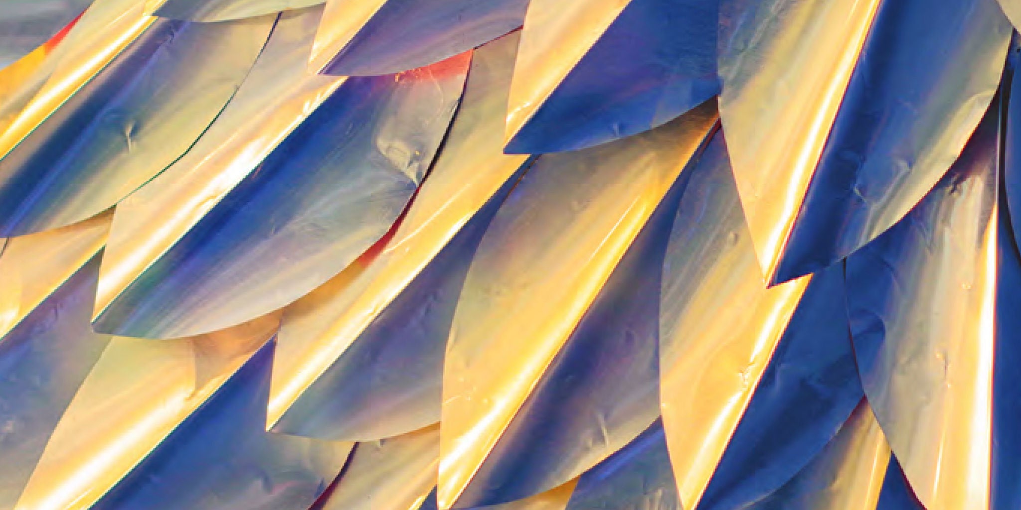 The image features a close-up view of overlapping, metallic, feather-like shapes arranged in a repetitive pattern. The feathers display an iridescent palette with hues of gold, blue, and purple, creating a dynamic and reflective visual effect. The abstract composition emphasizes texture and light, evoking a modern and artistic mood.