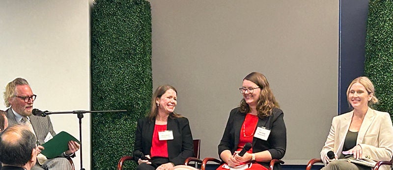 Four professionals sit at the front of a room engaged in a panel discussion. Three women and one man are seated on chairs, holding microphones and notes while speaking to an audience. They wear business attire and conference name badges, suggesting a formal event or seminar. The background features green hedge panels and a neutral wall, creating a modern corporate setting.