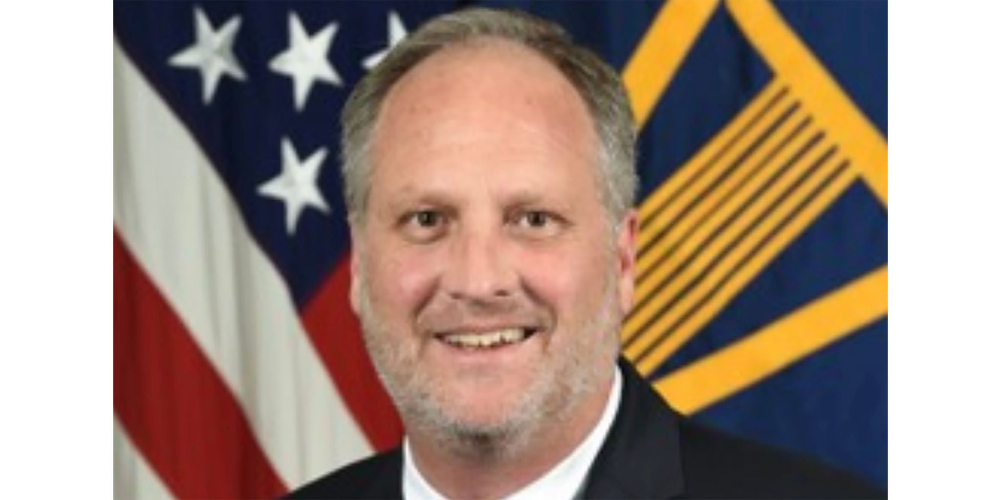 A man in formal attire is positioned in front of a backdrop featuring the United States flag and another flag with a geometric design. The setting suggests an official or governmental context, emphasizing professionalism and authority.