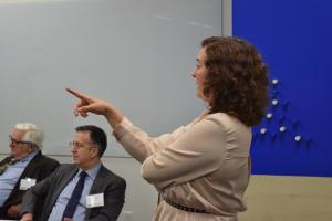 A woman stands and gestures with her hand while speaking in a professional meeting. Two seated men in business attire listen in the background. The setting appears to be a conference or seminar room with a whiteboard and a blue wall. The scene suggests an engaged discussion or presentation in a corporate environment.