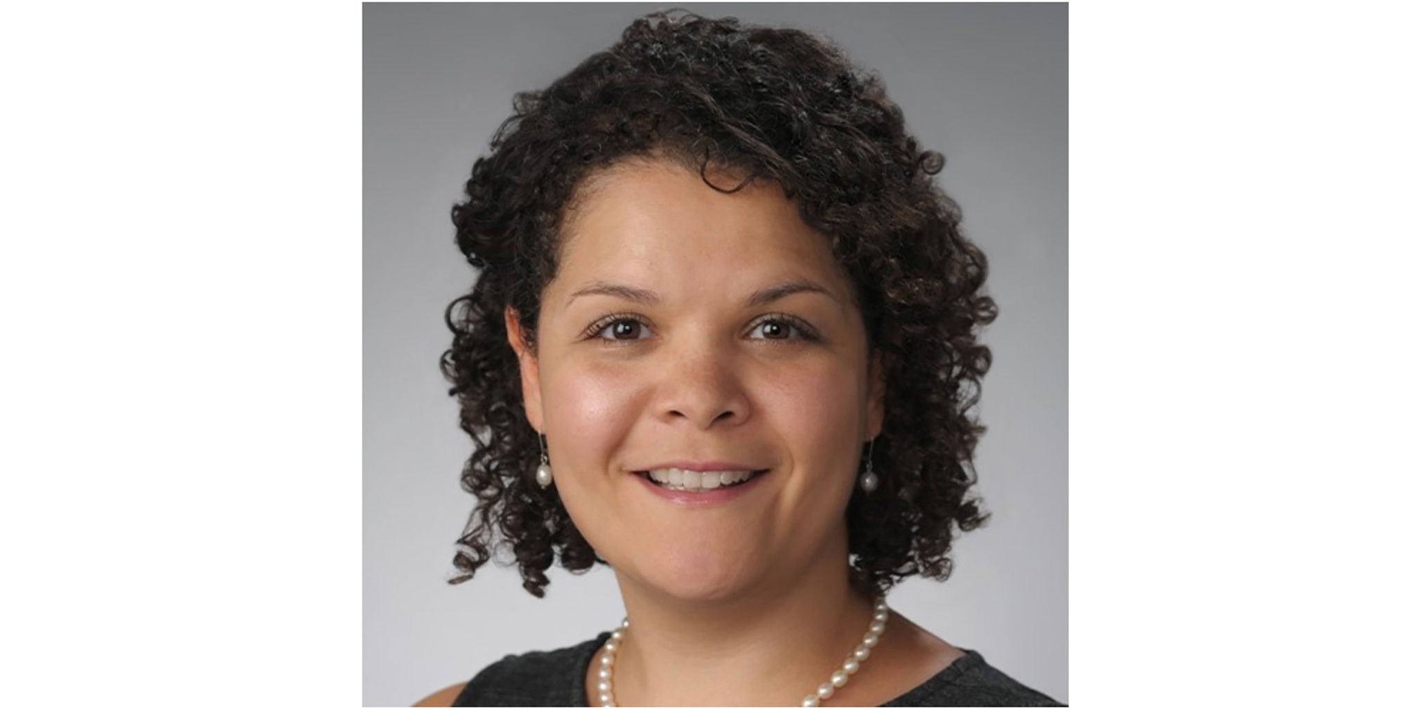 A professional portrait featuring a person wearing a black top and a pearl necklace. The background is a neutral gray, creating a formal and polished look. The subject's curly hair adds texture and character to the composition.