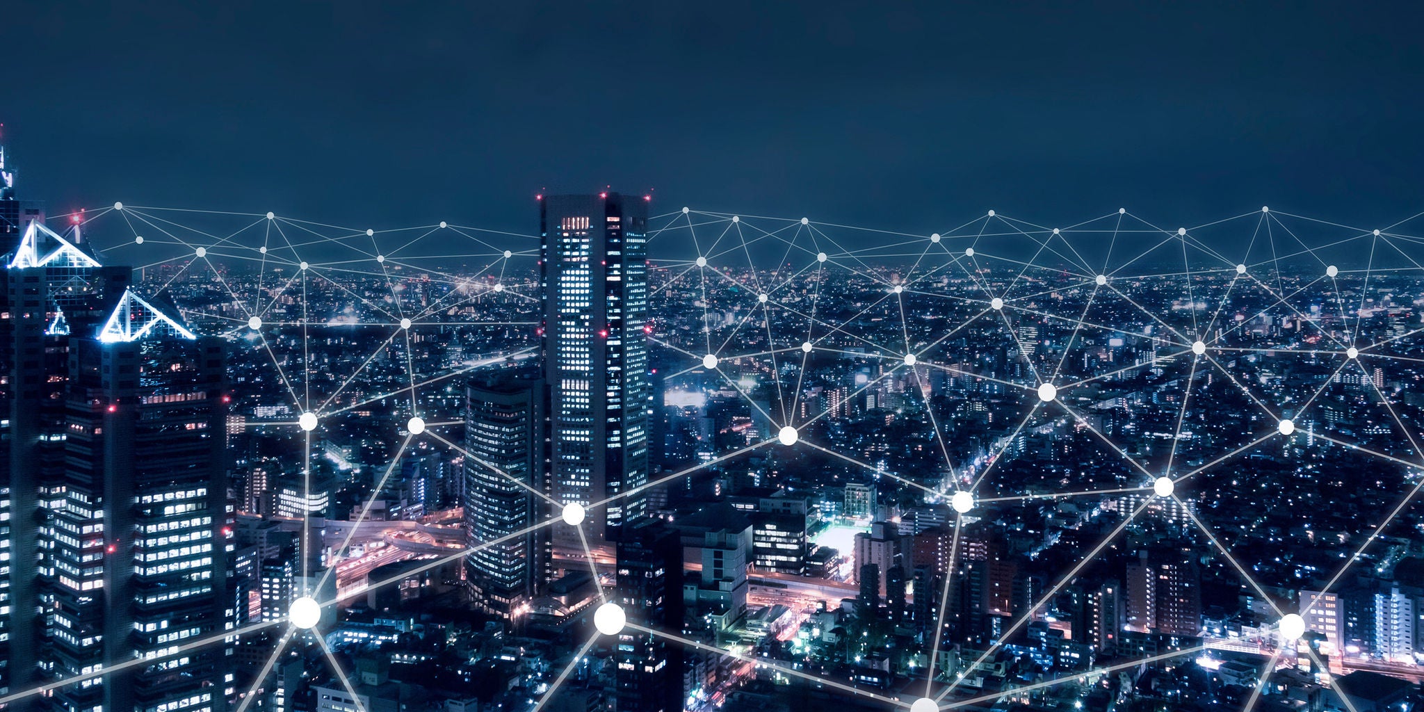 A modern city skyline is illuminated at night, featuring tall skyscrapers and dense urban buildings. A digital network overlay with glowing connection points and lines spans across the cityscape, symbolizing connectivity and smart technology. The overall mood is futuristic and technological, with a cool blue palette dominating the scene. No visible text or numbers are present in the image.