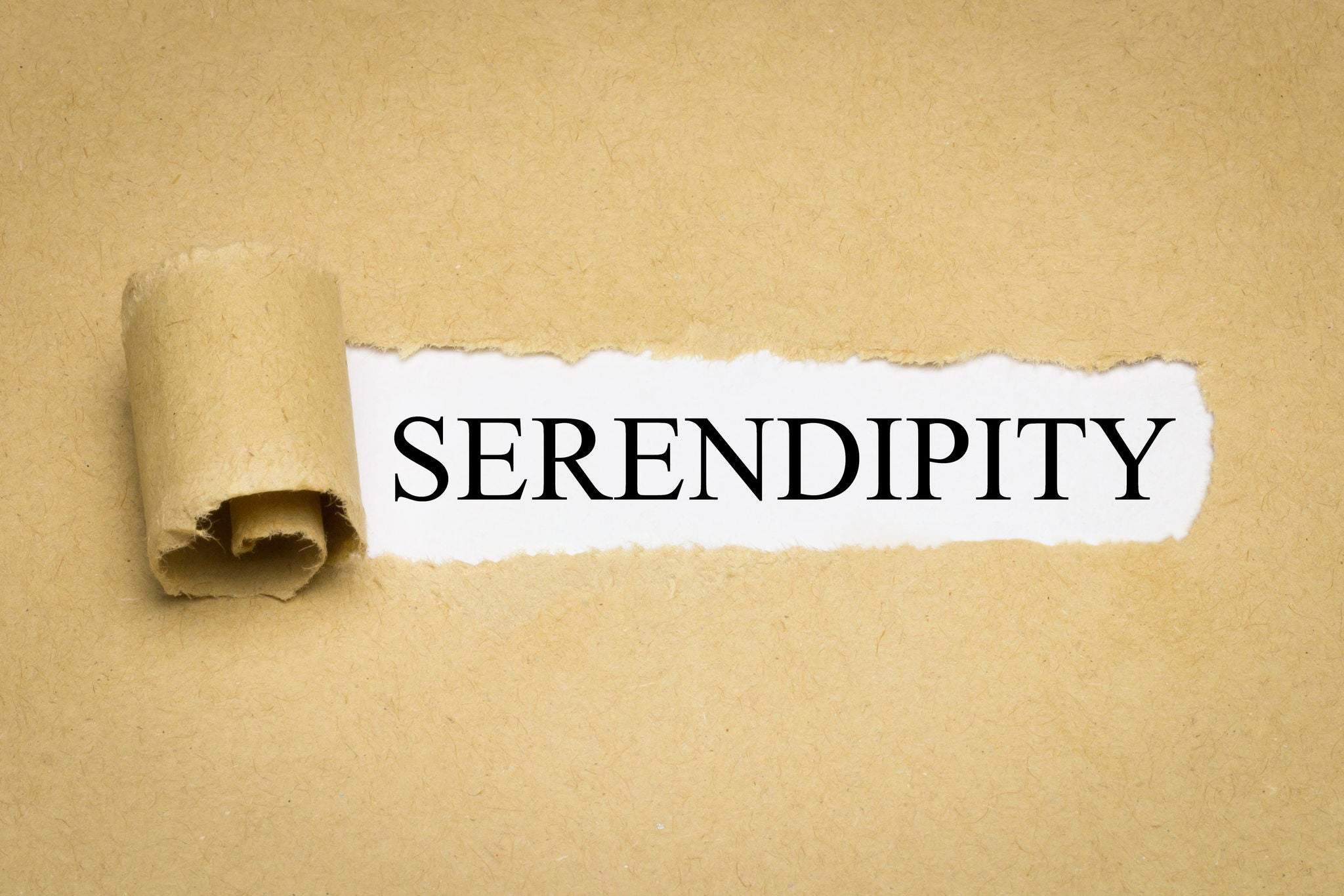 The word 'Serendipity' is prominently displayed in bold black text, revealed through a torn section of brown kraft paper. The composition emphasizes the concept of discovery and surprise. The neutral tones of the paper create a minimalist and elegant visual style.