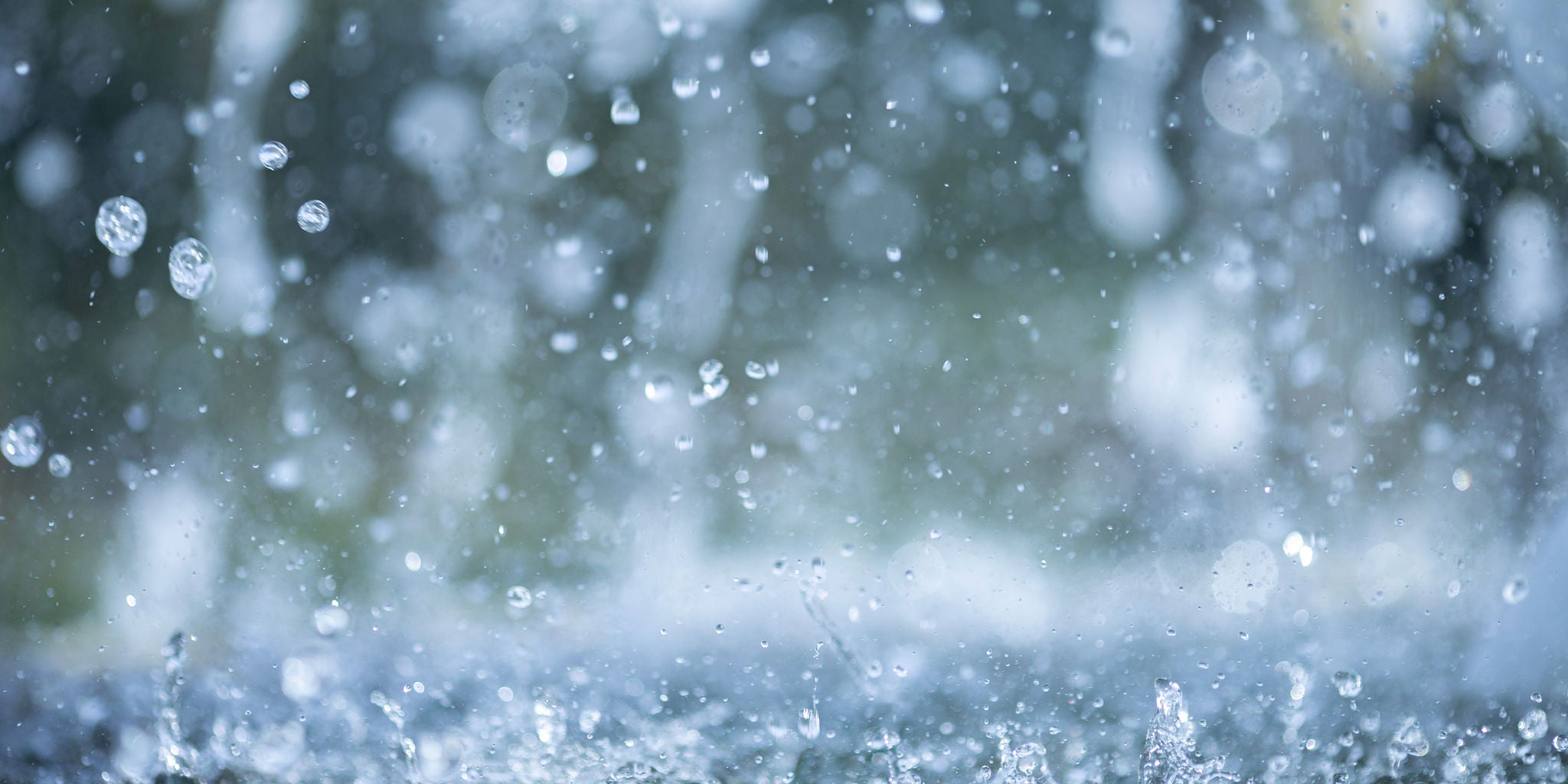 A detailed view of raindrops splashing on a wet surface, creating dynamic water droplets in motion. The background is softly blurred, emphasizing the movement and texture of the water. The scene conveys a fresh and natural atmosphere, with a cool color palette dominated by blues and grays.