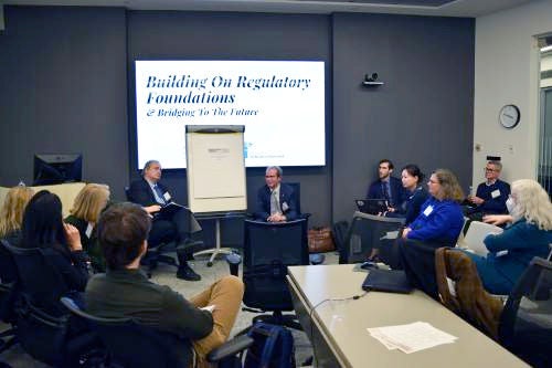 A group of professionals is seated in a modern conference room, participating in a panel discussion. The main screen displays the text 'Building On Regulatory Foundations & Bridging To The Future.' Two speakers are seated at the front, while attendees listen attentively. The setting is formal, with a collaborative and focused atmosphere.