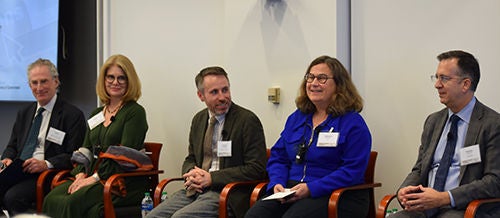 A group of five professionals sit in a row on chairs during a panel discussion. They wear business attire and conference name badges, suggesting a formal corporate or academic event. The setting appears to be an indoor conference room with neutral walls and a presentation screen partially visible on the side. The panelists look engaged and attentive, indicating an organized discussion or presentation.