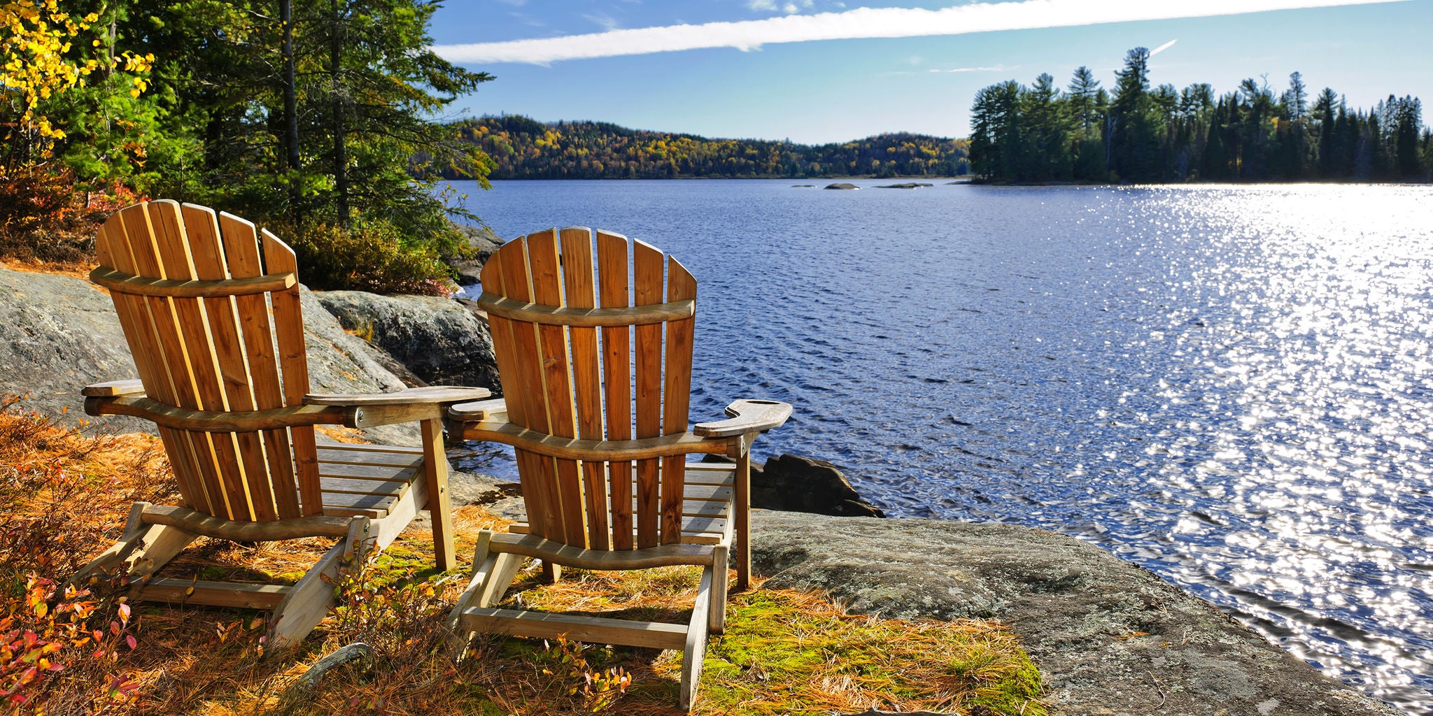 Two wooden Adirondack chairs are positioned on a rocky shoreline, facing a calm lake surrounded by forest. The scene is bathed in warm sunlight, with autumn foliage visible on the trees. The tranquil water and clear blue sky create a serene and inviting atmosphere. No people are present, emphasizing solitude and relaxation in nature.