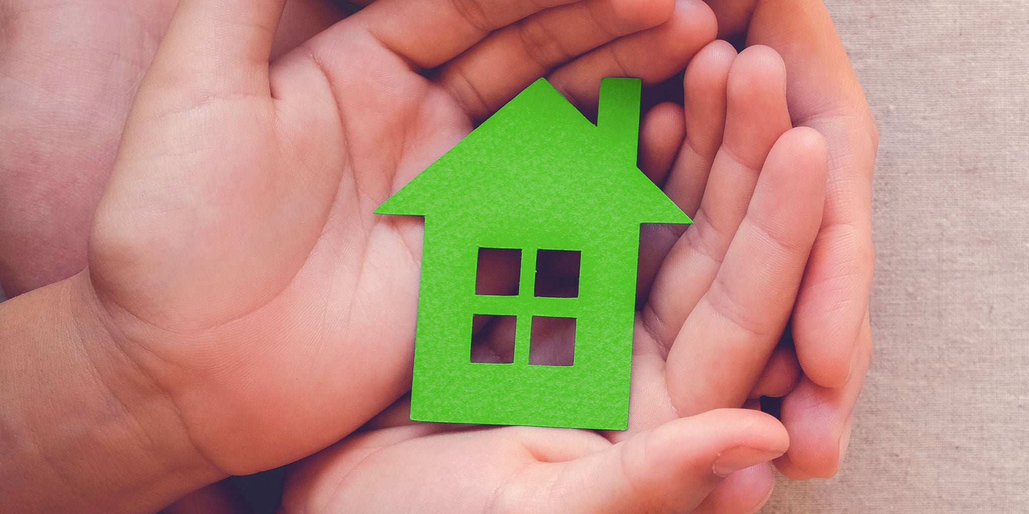 A pair of adult and child hands gently cradle a green paper house cutout. The scene suggests themes of family, safety, and home security. The background is neutral, focusing attention on the hands and the symbolic house. The image conveys warmth, care, and protection.