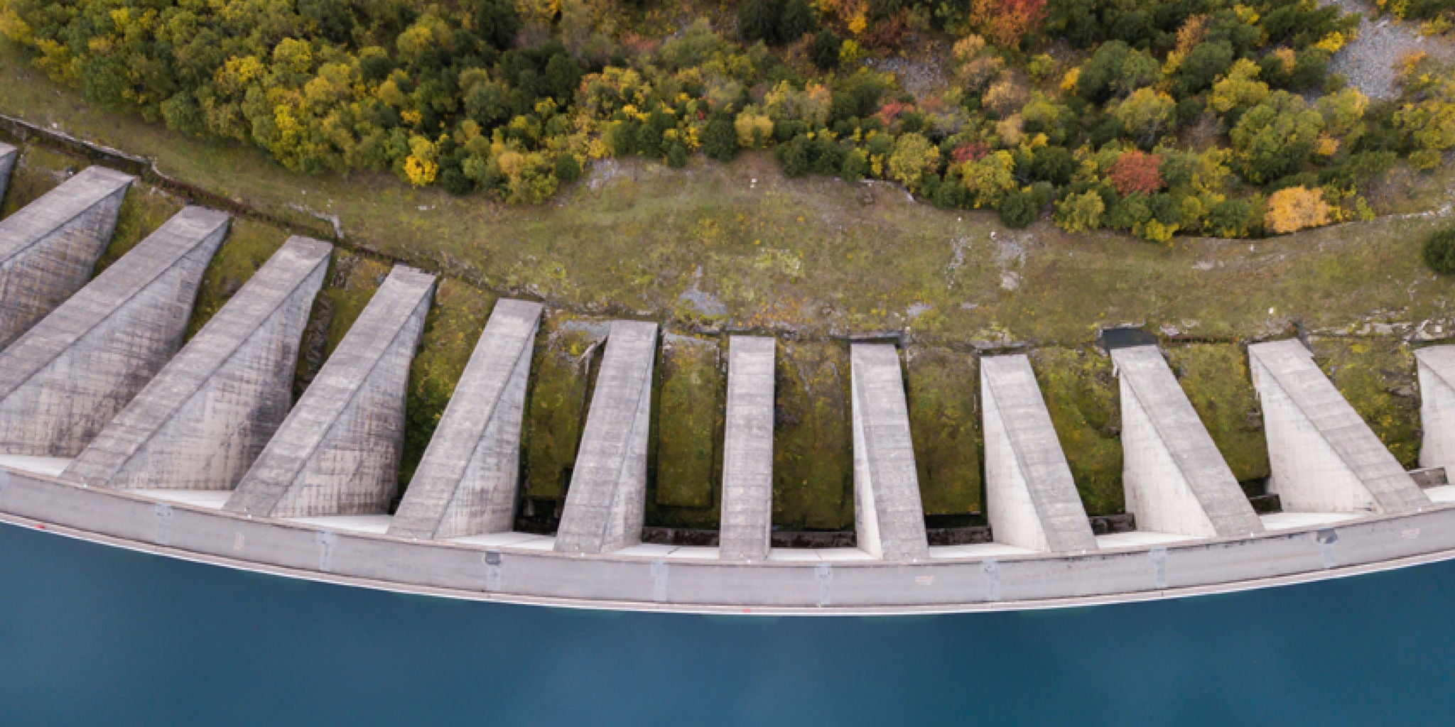 This image shows a top-down perspective of a large concrete dam holding back a body of blue water. The dam's geometric structure is clearly visible, with evenly spaced supports casting shadows. Behind the dam, a strip of green and yellow foliage lines the landscape, indicating a natural setting in early autumn. No visible text, numbers, or people are present in the image.