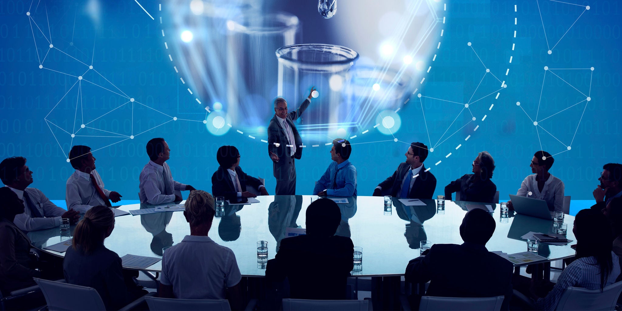 A group of professionals sits around a large conference table, attentively watching a presenter who points to a digital screen. The background features scientific visuals, including laboratory glassware and molecular structures, set against a blue, high-tech interface. The scene conveys innovation and collaboration in a modern corporate environment. The group includes both men and women of diverse ages, dressed in business attire.