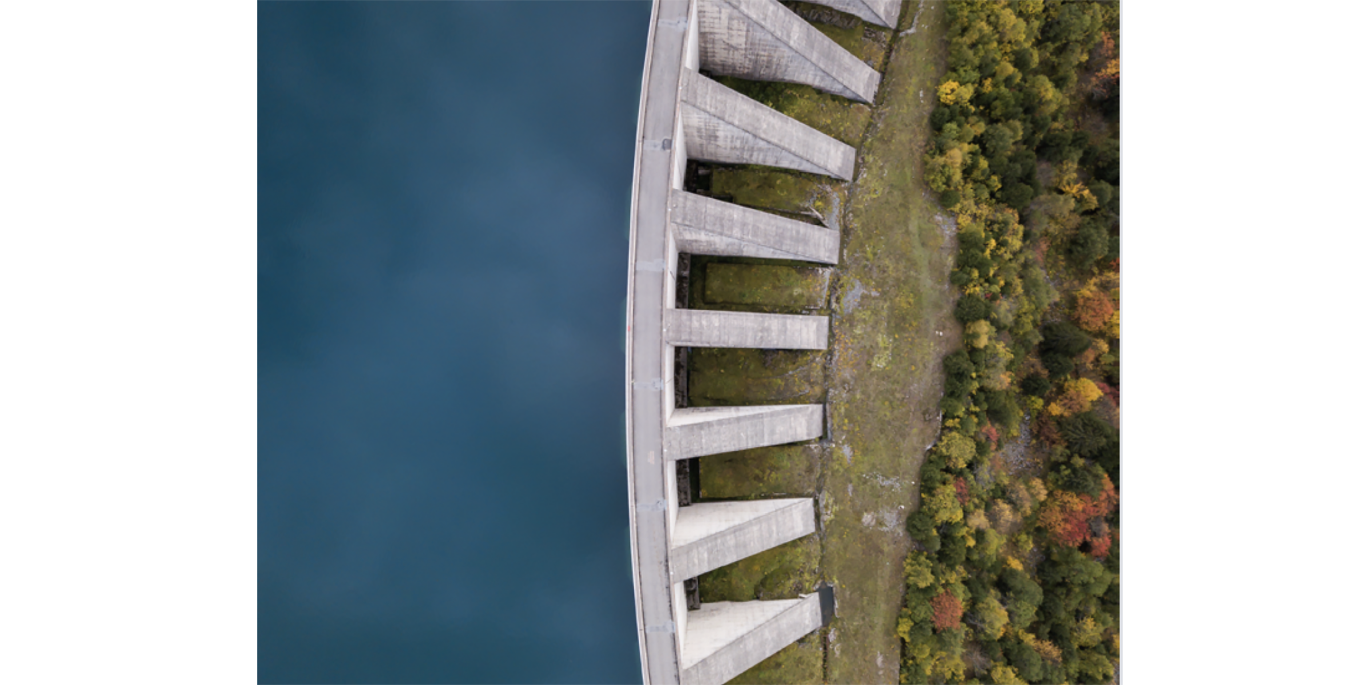 An aerial perspective of a massive concrete dam spanning across a body of water. The structure is surrounded by lush greenery and autumn-colored trees on one side. The calm blue water contrasts with the vibrant natural landscape, creating a striking visual balance.