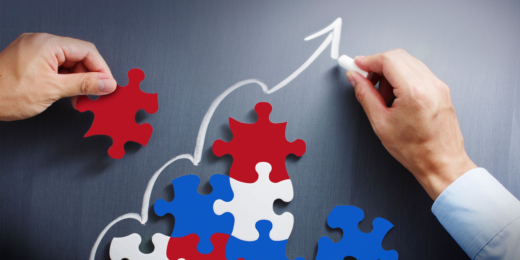 Two hands are seen placing red, blue, and white puzzle pieces together on a dark surface. An upward-pointing arrow is drawn in white chalk, symbolizing growth or progress. The scene suggests teamwork, strategy, and goal achievement. The overall mood is optimistic and professional, with a focus on collaboration.