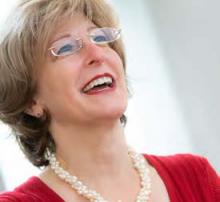 A middle-aged woman smiles brightly while looking slightly upward. She is wearing glasses, a red top, and a white beaded necklace. The background is softly blurred, suggesting an indoor setting with natural light. The overall mood is cheerful and positive.