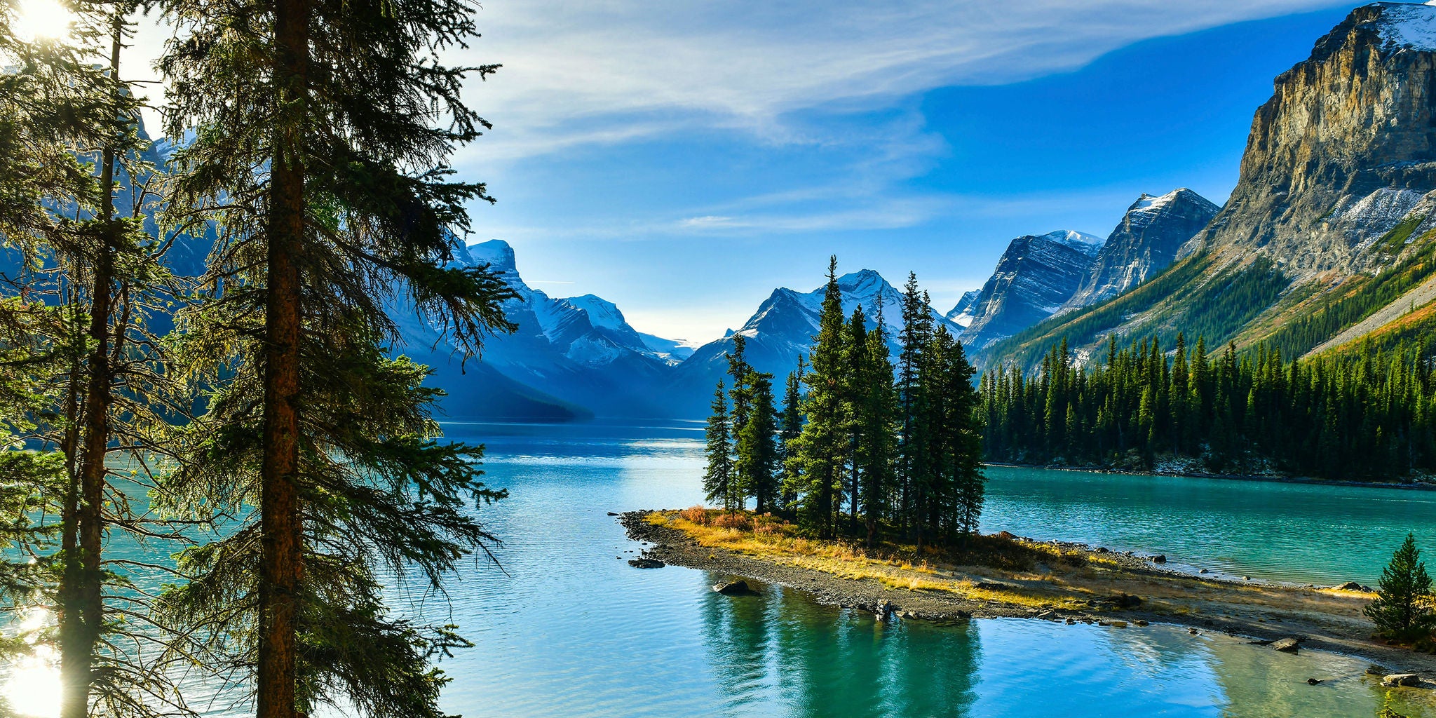 A tranquil mountain lake features a small island covered with tall pine trees, surrounded by clear blue water. Snow-capped peaks rise in the background under a vivid blue sky with wispy clouds. The scene is bathed in bright daylight, highlighting the lush greenery and rugged terrain. No people or visible text are present, emphasizing the untouched natural beauty. The overall mood is peaceful and refreshing.