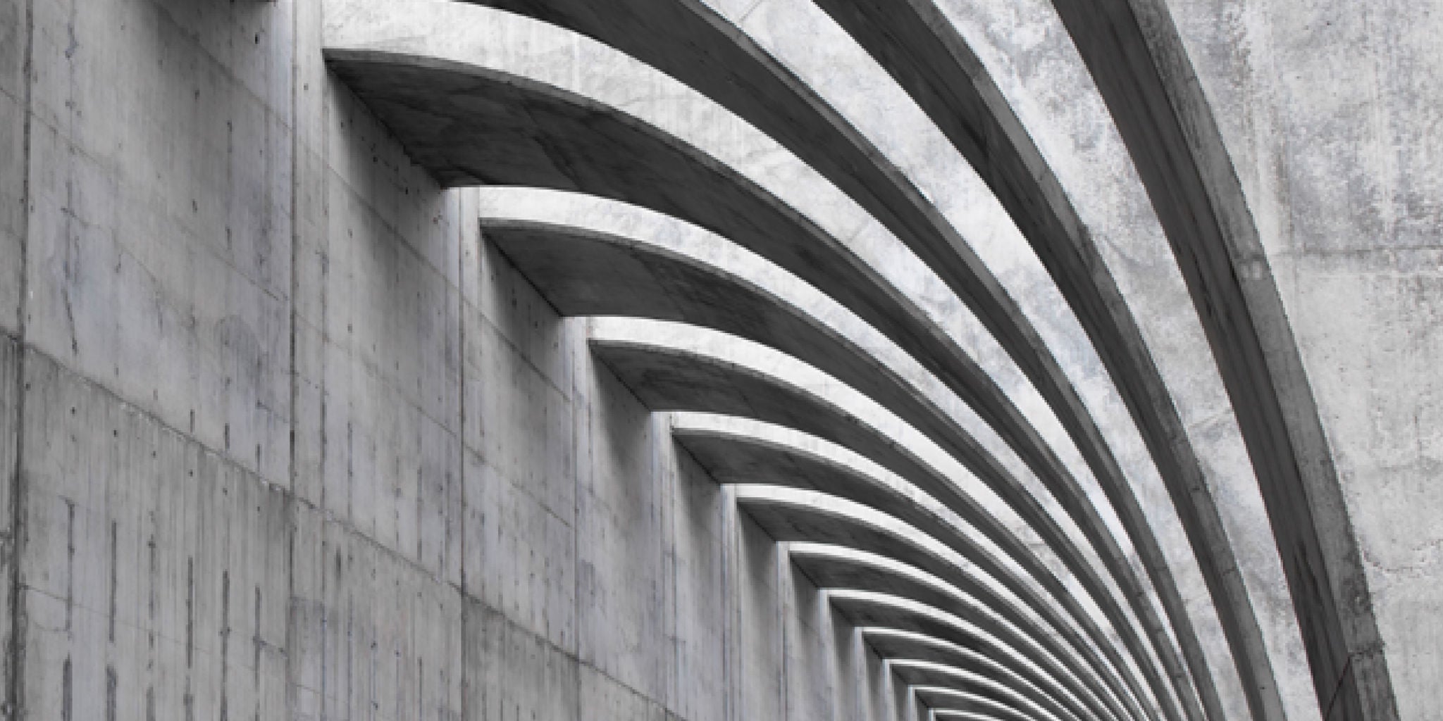 The image features a series of repeating concrete arches forming a striking architectural pattern. The setting appears to be an interior or exterior of a modern building, emphasizing geometric lines and symmetry. The color palette is dominated by shades of gray, contributing to a minimalist and industrial mood. No people, text, or numbers are visible in the image.