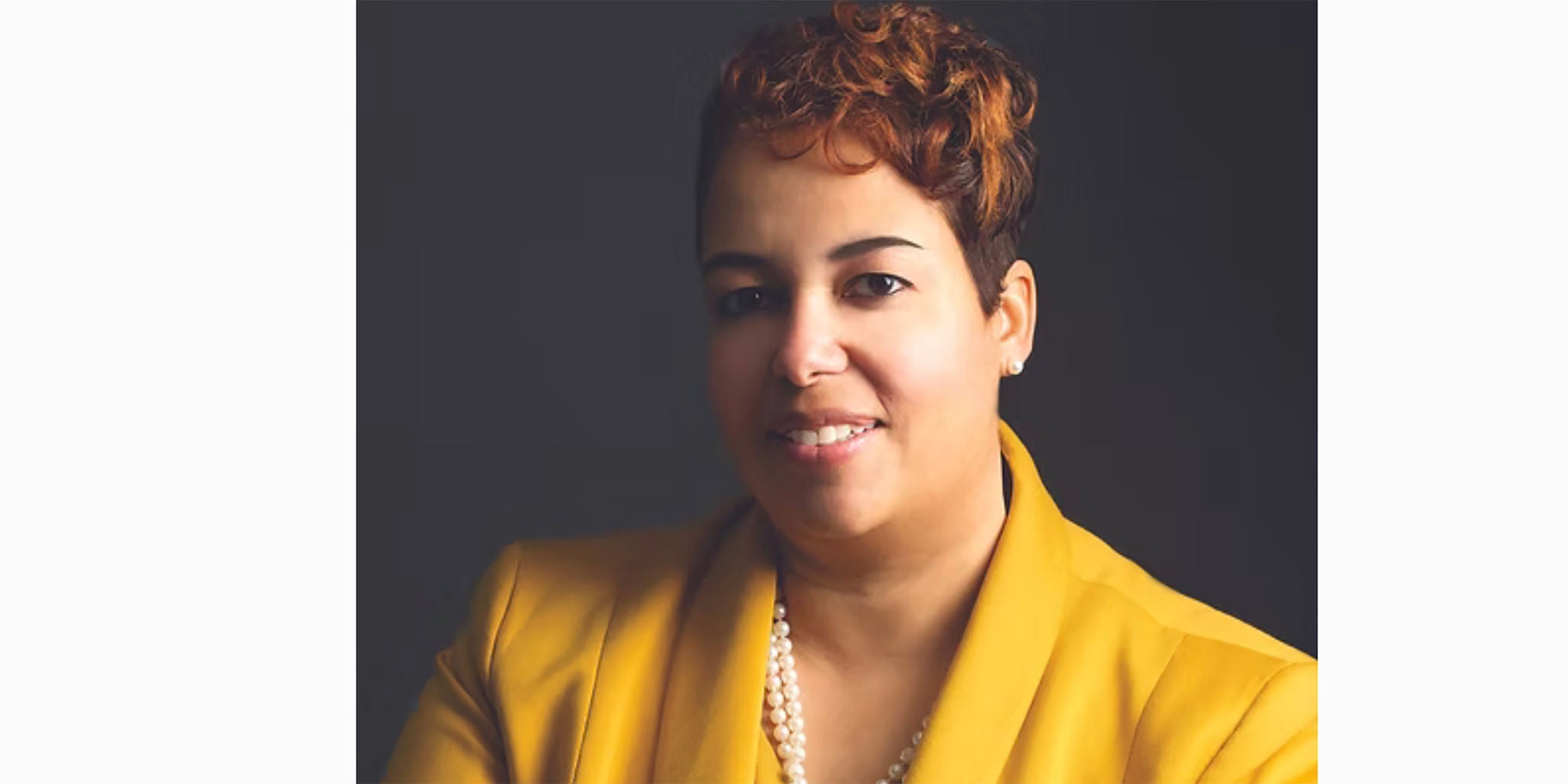 A person wearing a bright yellow blazer paired with a pearl necklace is set against a dark background. The outfit exudes a professional and stylish vibe. The lighting highlights the vibrant color of the blazer and the texture of the pearls.