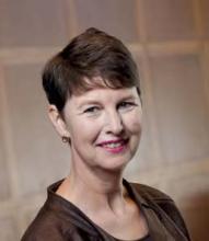 A middle-aged woman is captured in a professional headshot-style portrait. She is smiling gently and looking toward the camera, suggesting a confident and approachable demeanor. The background is softly blurred with warm, neutral tones, drawing focus to her face. The overall setting and lighting create a polished, corporate or business-like atmosphere.