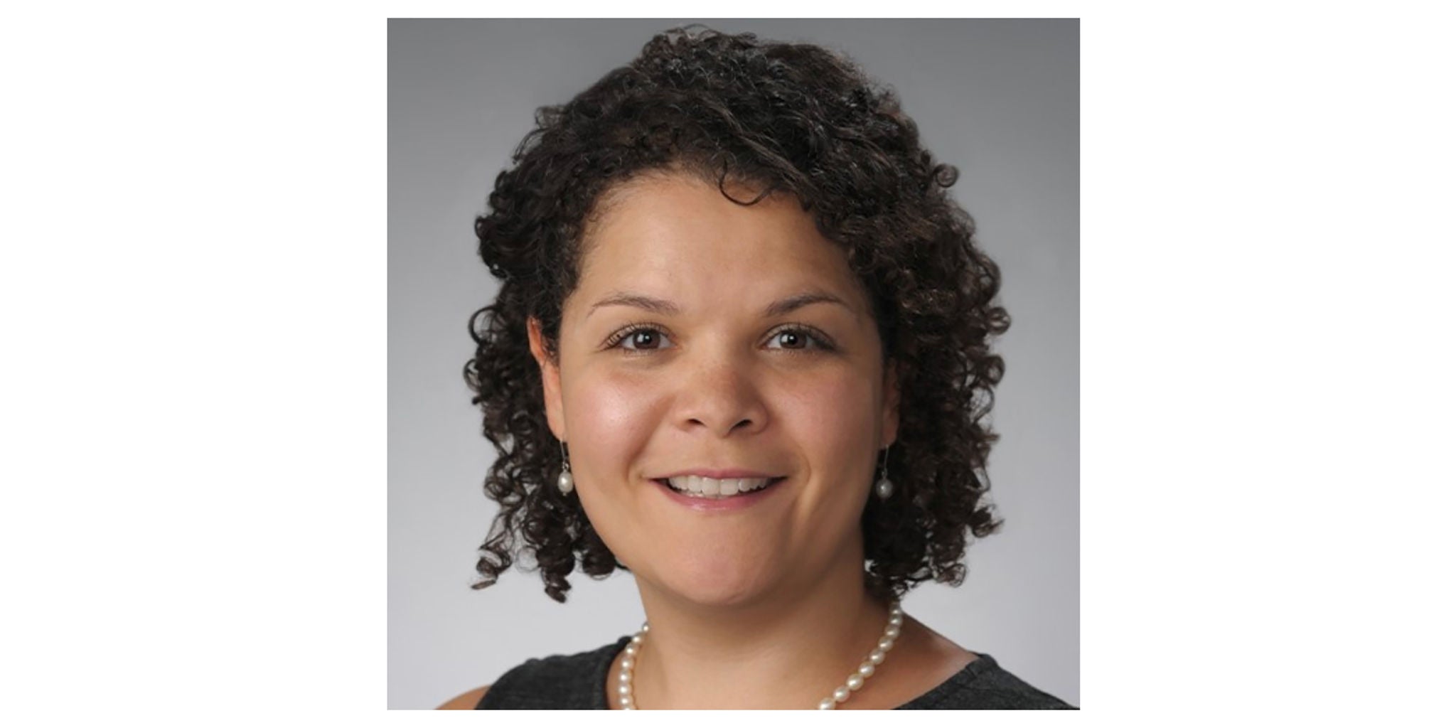 A woman with curly hair is seen wearing a pearl necklace and a sleeveless black top. The background is a neutral gray, creating a professional and formal setting. The overall mood is elegant and sophisticated.