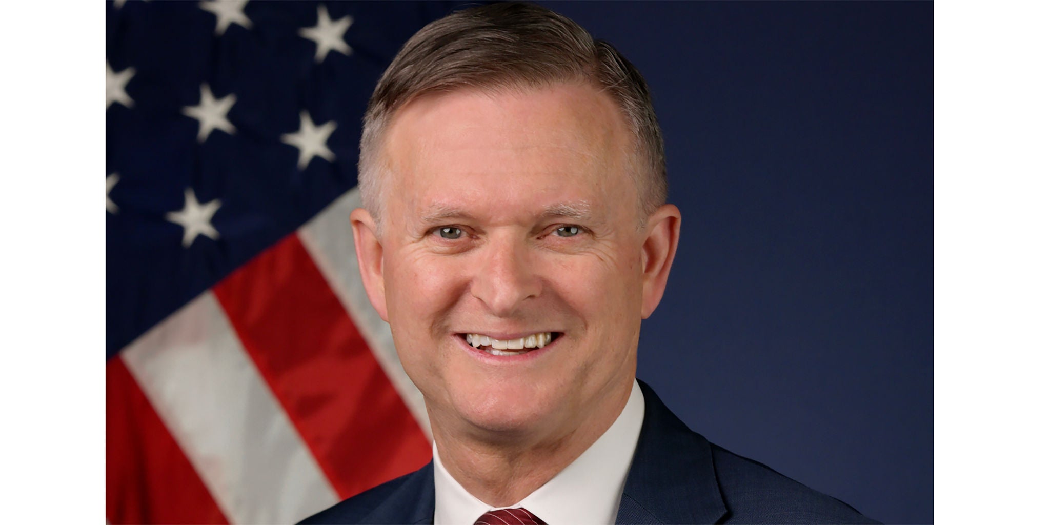 A professional portrait featuring a person in formal attire, set against a backdrop of the American flag. The image conveys a patriotic and official tone, with the flag prominently displayed in the background. The subject is dressed in a suit and tie, emphasizing formality and professionalism.