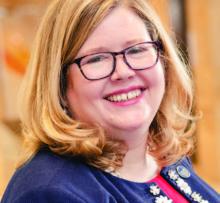 A woman with shoulder-length blonde hair and glasses smiles warmly at the camera. She is wearing a navy jacket over a red top, accessorized with a decorative necklace. The background is softly blurred, suggesting an indoor professional or office environment. The overall scene conveys a friendly, approachable, and professional mood.
