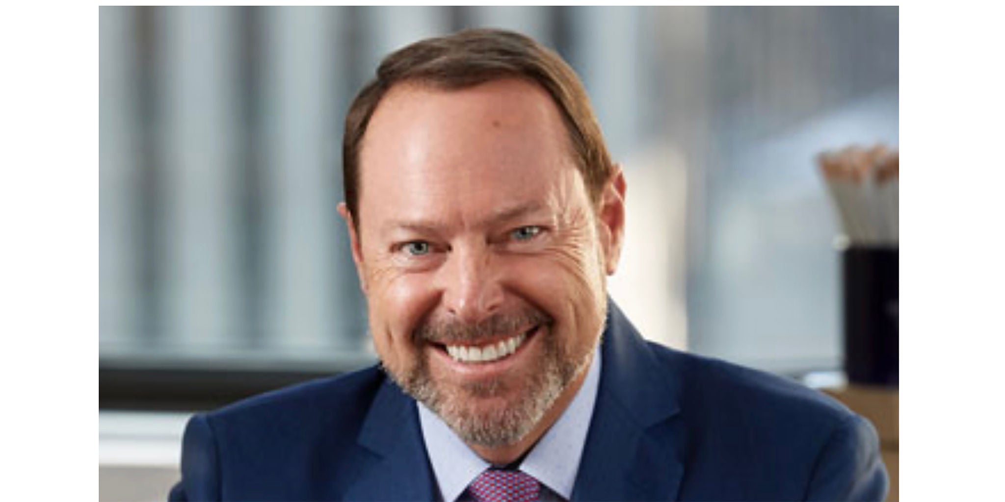 A person wearing a blue suit and pink tie is seated in a professional office environment. The background features blurred windows and office supplies, suggesting a corporate or business setting. The overall tone is formal and professional.