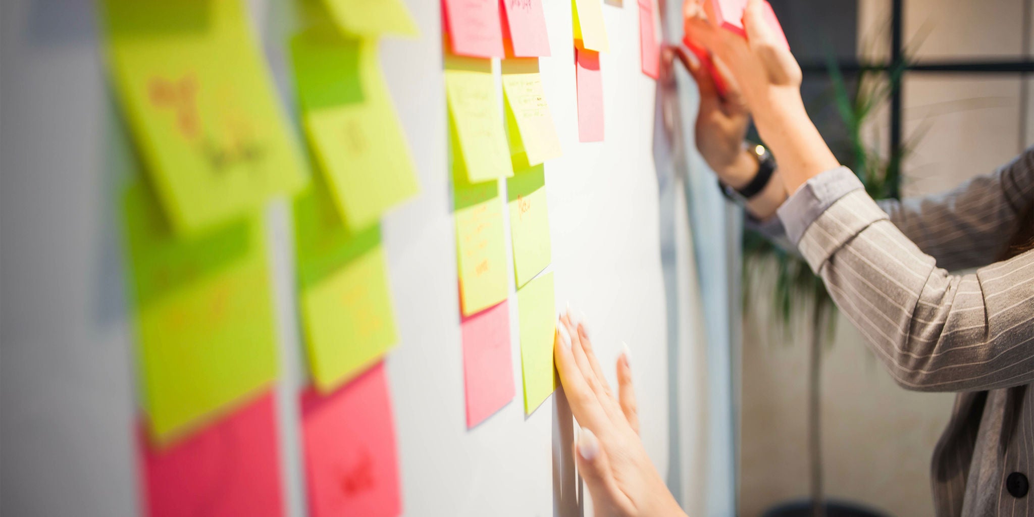 A collaborative team activity featuring individuals placing colorful sticky notes on a whiteboard. The setting appears to be an office or creative workspace, fostering teamwork and idea generation. Bright pink, yellow, and green notes dominate the visual palette.
